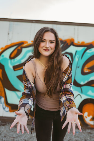 Senior girl wearing a tan tank top and flannel shirt with black jeans smiles at camera in downtown urban setting at sunset