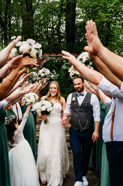A bride and groom smiling as they walk through a tunnel of hands that their bridesmaids and groomsmen created