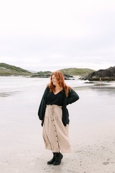 Lelia Angelique standing on a serene beach with a peaceful expression, wearing a cozy sweater and long skirt, with scenic hills in the background.