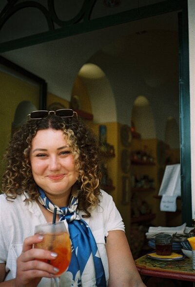 Andrea Cakmar sits at a café, smiling warmly at the camera while holding a drink. She is wearing a white top with a blue scarf and sunglasses perched on her head.