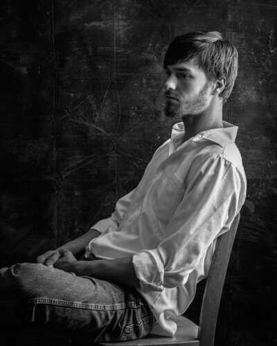 black and white image of man sitting in a chair in jeans and white button down shirt with dramatic lighting