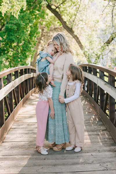 A mom and her three daughters hold hands and snuggle on a wooden bridge