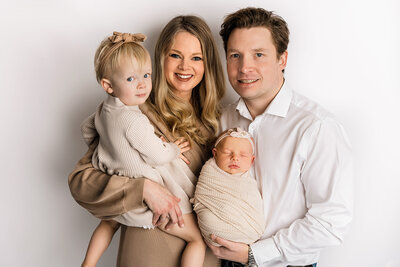 Happy family of four portrait with parents, toddler daughter, and sleeping newborn baby in coordinating neutral tones photographed in studio