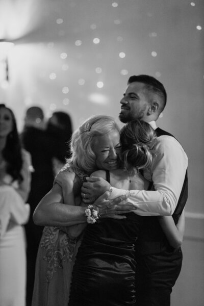 The groom hugging his mom and daughter as they dance at the reception, in black and white