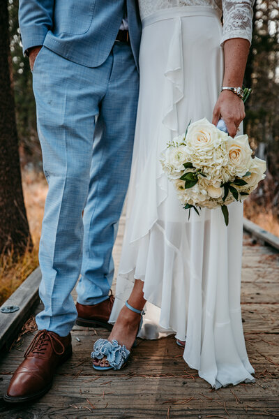Close-up of the bride’s bouquet and shoes alongside the groom’s suit, standing on a wooden bridge during their elopement.