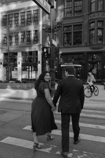 A couple holding hands and walking across the street as one looks back over their shoulder 
