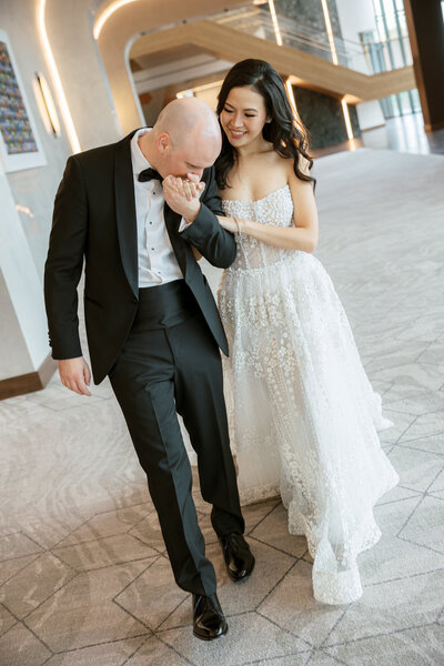 Bride and groom walking together down a modern, well-lit hallway. The bride wears a sparkling, off-the-shoulder gown, and the groom is in a classic black tuxedo. They smile at each other, holding hands.
