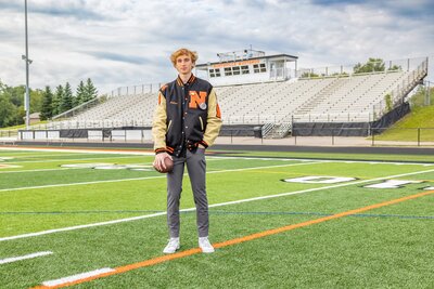 A male high school senior is standing on a football field wearing a varsity jacket and holding a football