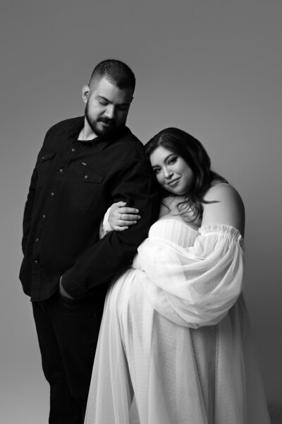 Black and white studio maternity portrait of expecting couple, woman in sheer gown leaning on partner, taken in Austin, Texas.