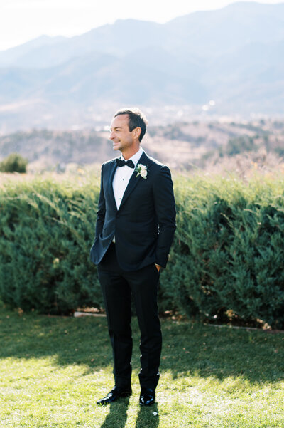Groom in tux with hands in pockets outside with green bushes behind him by Colorado Wedding Photographer JKG Photography
