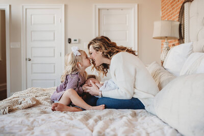 Mother in white sweater with newborn on bed with blonde daughter and a white bow in her hair