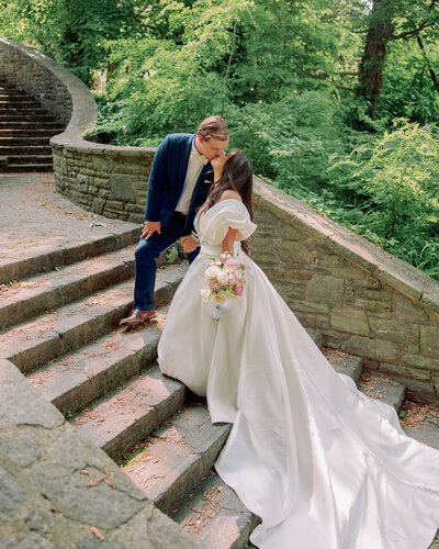 A newlywed leaning down and kissing their partner as they walk up a stone staircase 