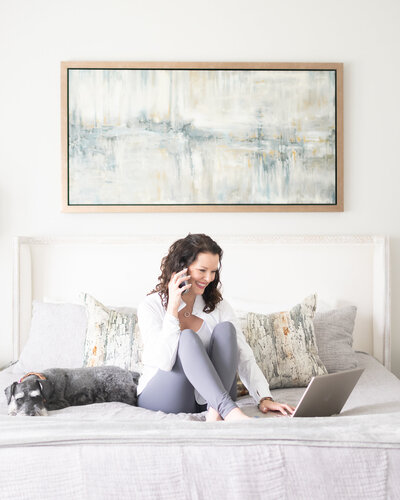woman in white jacket and grey pants on phone and computer in master bedroom