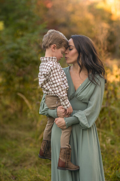 mother in sage green dress holding toddler son in beige pants brow boots and white and brown plaid shirt touching foreheads in the fall foliage of cleveland ohio