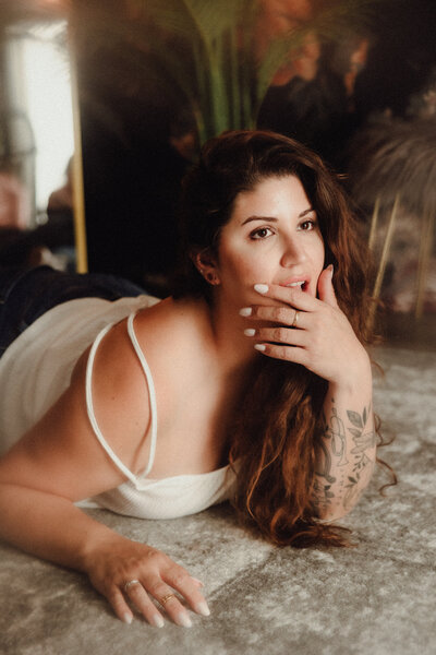 Woman with tattooed arm lays on the floor, gently touching her lips and gazing toward the camera in a dreamy boudoir setting.
