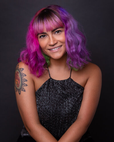 colorful-haired woman in a black sequence top seated in front of a black background with both hand lightly crossed