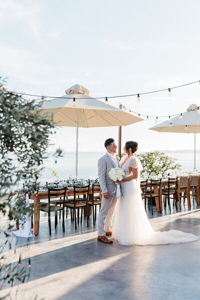 Bride and groom share a quiet moment beside their waterfront reception setup with string lights and parasols, overlooking the sea at their destination wedding in Greece