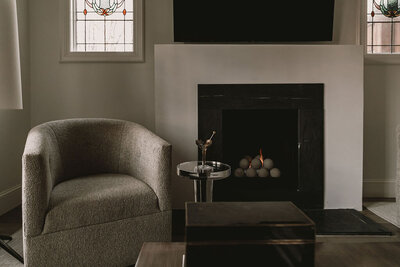 A modern living room with a gray armchair, a round side table, and a fireplace. A television is mounted above the fireplace, and stained glass windows are on both sides. Perfect for professionals who hire an online business manager to streamline their needs without compromising on comfort.