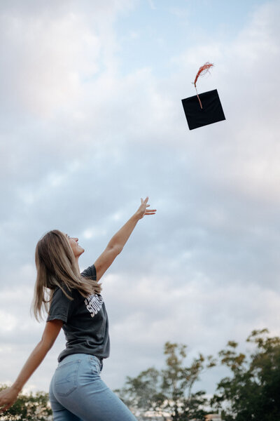 A graduating senior female in a casual University of South Carolina t-shirt/jeans throws her graduation cap in the air. The photo is angled so you can see the blue South Carolina sky.