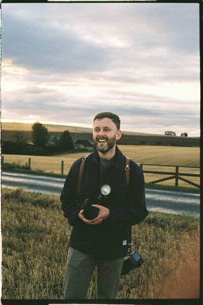 Aberdeen wedding photographer Scott Arlow stands in a field holding a camera.