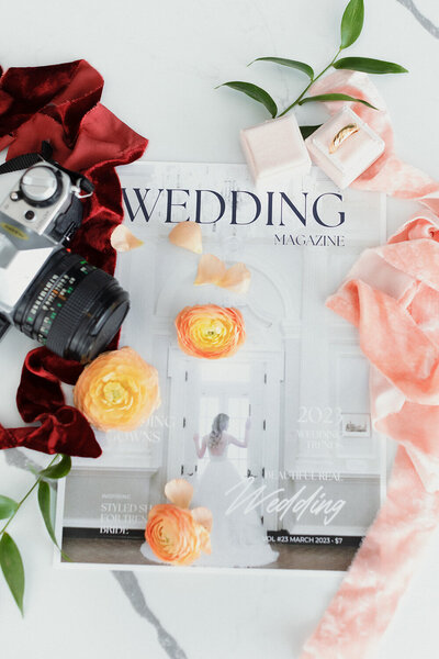 A wedding magazine surrounded by a Canon camera, red and pink cloth, flowers, leaves and a wedding ring