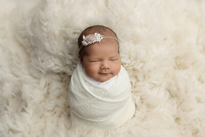 A newborn baby sleeps with a smile in a white swaddle and pink flower headband