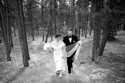 A bride and groom walk up a path among trees as the groom holds the bride's veil and train.