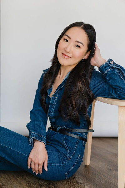 Woman in denim jumpsuit leans against a wooden stool while sitting on the floor of a photo studio in North Carolina | Sara Coffin Photo