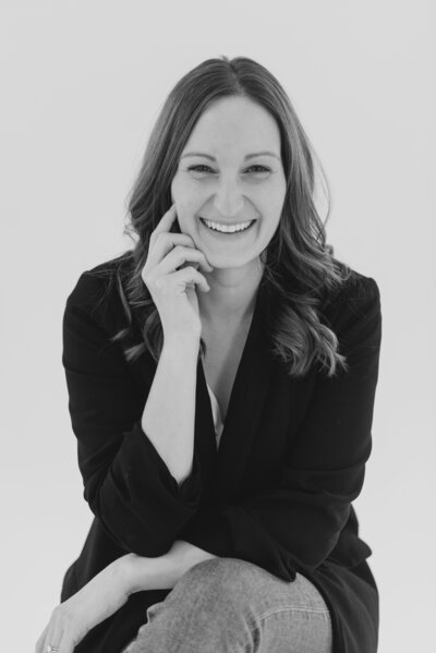 Columbus headshots of brand photographer sitting on a white backdrop with her legs crossed and her elbows resting on her knees as she smiles