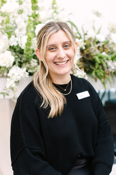 Kelsey Schultz, event sales coordinator at SoHo63, a Chandler wedding and event venue, smiles in front of a floral backdrop. Her approachable style and attention to detail help make every SoHo63 wedding and event seamless and memorable.