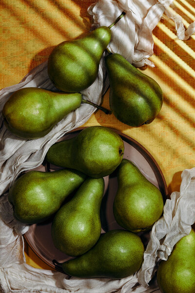 A serene still-life image featuring green pears arranged on a plate, bathed in soft natural light. Photographed by Mica McCook, this image captures the organic beauty and rich textures of fresh fruit against a warm, textured backdrop.