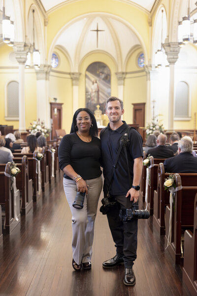 Two photographers pose inside a church with yellow walls and an ornate altar. Both hold cameras; the woman stands with her hands on her hips while the man has an arm around her waist. The pews are filled with people, and floral decorations adorn the aisles.