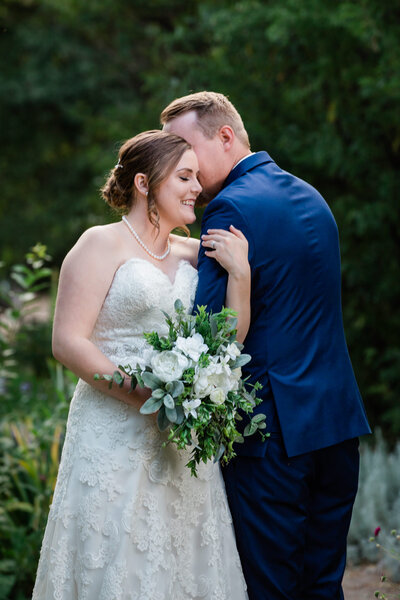 A husband wearing a blue suit whispers in his new bride's ear while she holds his arm, closes her eyes, and smiles.