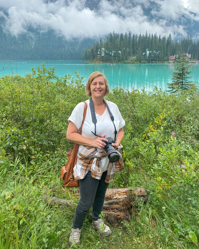 Woman with camera around her neck standing in front of a glacier blue lake with fog rolling above.