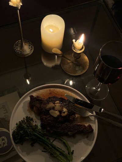 Romantic dinner setting with a plate of grilled steak topped with garlic, steamed broccolini, red wine glass and glowing candles on a dark table