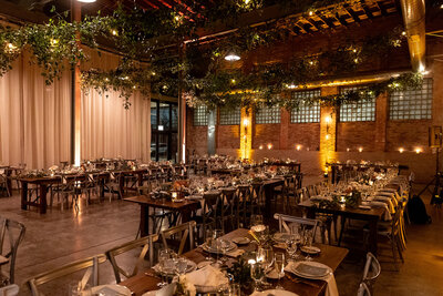 Warmly lit rustic dining hall with foliage draped from the ceiling and elegant, minimalistic table decor.
