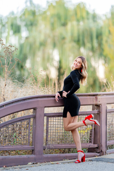 girl in black dress with red heels kicking one foot behind her and smiling in forest park