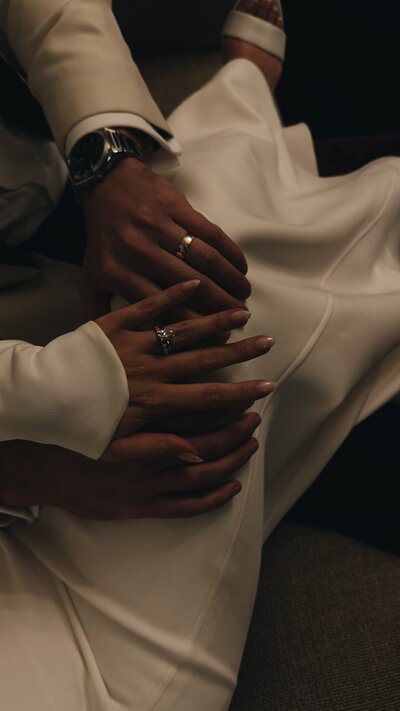 Hands resting on a white dress, adorned with wedding rings. The mood is intimate and tender, highlighting a special romantic moment.