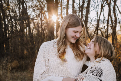Two people smiling at each other with knit shawls in a forest during sunset.