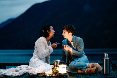 a couple in their wedding attire share a cup of tea at the edge of a lake, they are lit by lanterns as they end their adventure elopment. 