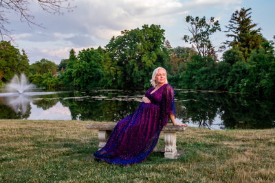 A pregnant woman sitting on a bench in front of a lake looks toward the sunset.