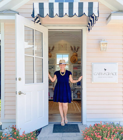 Founder standing in the doorway of the Gaby & Grace retail boutique in Galveston’s Evia community, smiling under a navy-and-white striped awning.