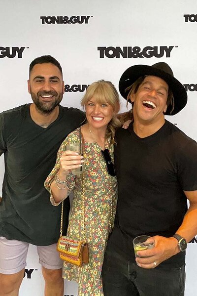 Two men and a woman posing with drinks in front of a branded Toni & Guy backdrop at a social or corporate event.