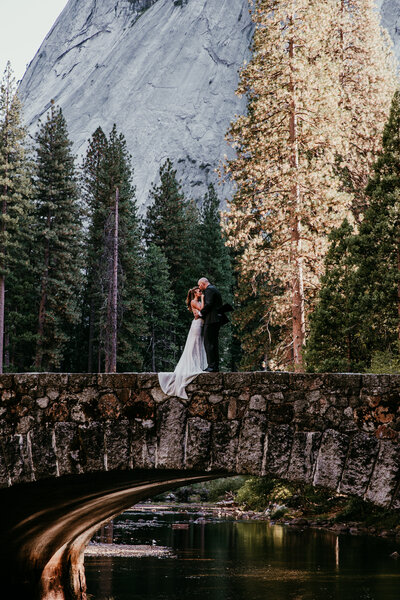 A bride and groom share a romantic kiss standing atop a historic stone bridge in Yosemite National Park, framed by towering pine trees and an impressive granite mountain backdrop.