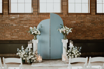 Wedding ceremony backdrop provided by Beautifully Layered Event Rentals in Milwaukee with 2 tall rounded light blue panel backdrops at staggered heights, white pillars and end tables holding white and blue floral bouquets and clear candle holders with a vintage looking rug and bouquets and vases on the floor surrounding. White chairs are set up for guests in front of it