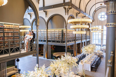 modern eclectic space with wine cellars along the wall, copper and brown surroundings, tall cieling with circle window and wedding reception rectangle tables covered in white wildflower like floral arrangements
