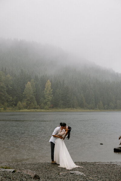 Bride and groom kiss at the edge of a still alpine lake in Oregon, their reflection visible in the water.