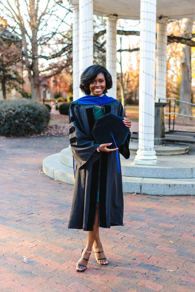 UNC Chapel Hill PhD graduate in gown and hood holding tam awhile standing in front of the Old Well.