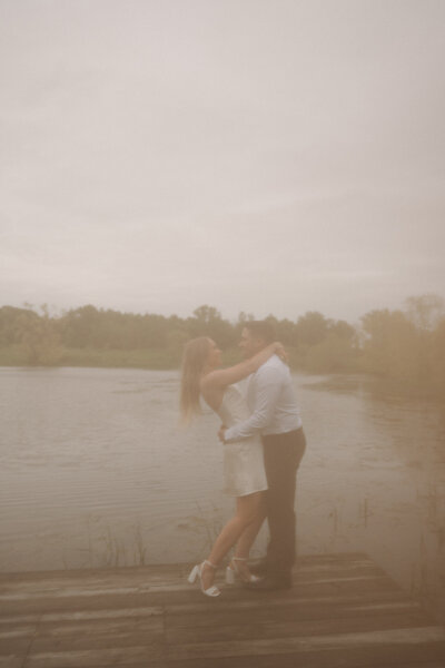 Engaged couple facing each other with arms draped over each other’s necks, sharing an intimate moment while overlooking a peaceful pond at Sterling Nature Center.