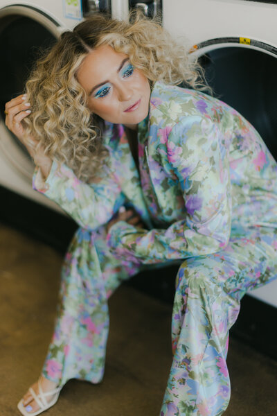 A stylish woman with voluminous curly blonde hair and vibrant blue eye makeup poses confidently in a pastel floral satin suit while crouched in front of a laundromat machine. Captured by Dani Lacey in Greenville SC, this bold branding image highlights vibrant fashion and fearless attitude.